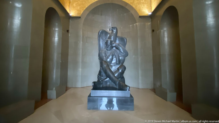 Seated dark Njegoš statue on a pedestal at the end of a pale arched chamber beneath a glowing gold-toned ceiling.