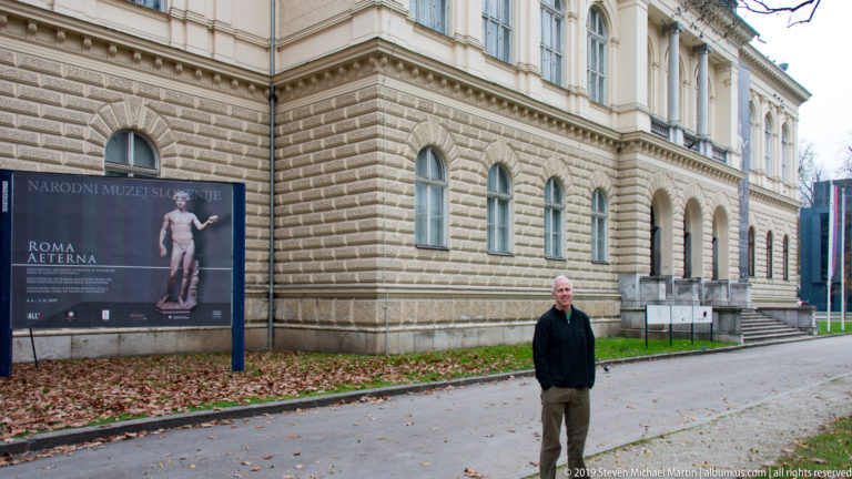 National Museum of Slovenia exterior with rusticated stone façade, arched windows, Roma Aeterna exhibition sign, and a man in front.