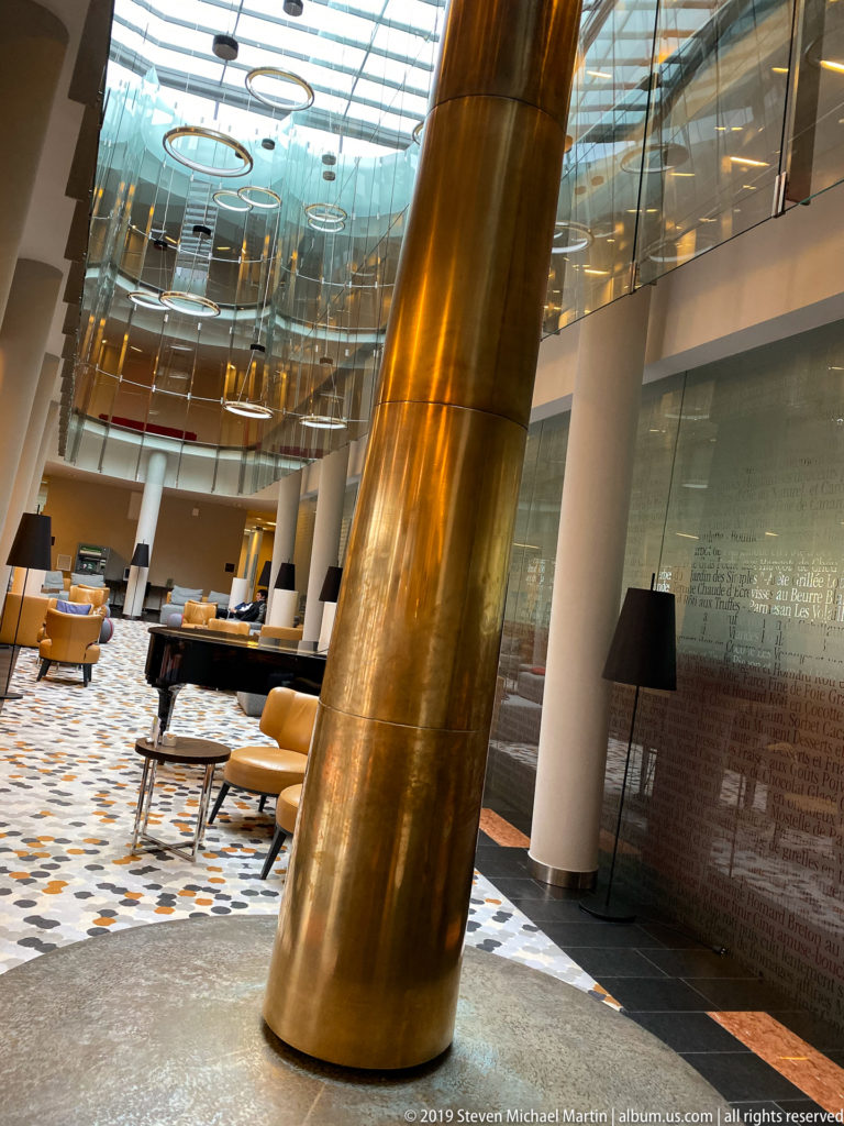 Contemporary hotel atrium with a massive gold-toned column, glass upper levels, patterned floor, lounge seating, and black lamps.