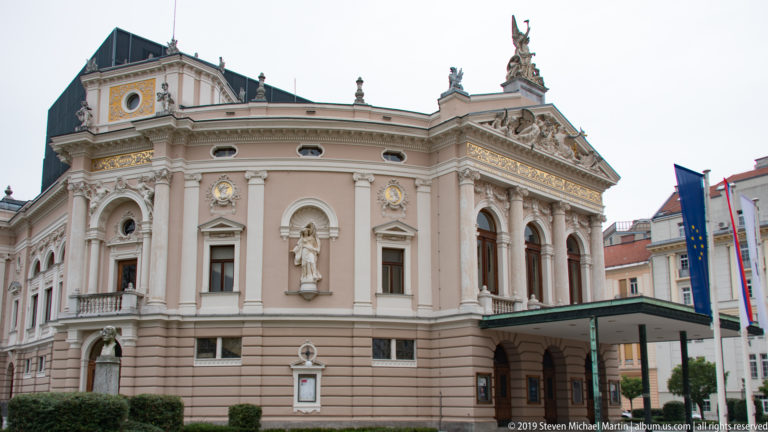 Oblique exterior view of Ljubljana’s opera house with pink façade, statuary, ornate pediment, and entrance canopy.