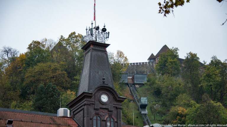 Dark ornamental clock tower in the foreground with Ljubljana Castle and its funicular track rising on the wooded hill behind.