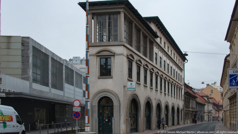 Historic multi-story building with arched windows and glazed upper pavilion beside a modern metal-clad structure in central Ljubljana.