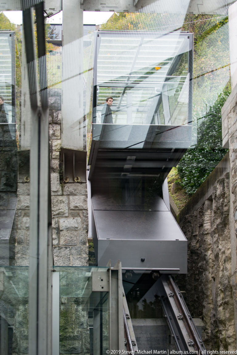 Glass-sided Ljubljana Castle funicular car on its steep track beside stone walls and modern station structure.