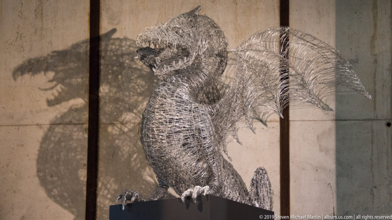 Wire-mesh bird sculpture on a dark pedestal, with a hooked beak, folded wings, talons, and a strong shadow on the wall.