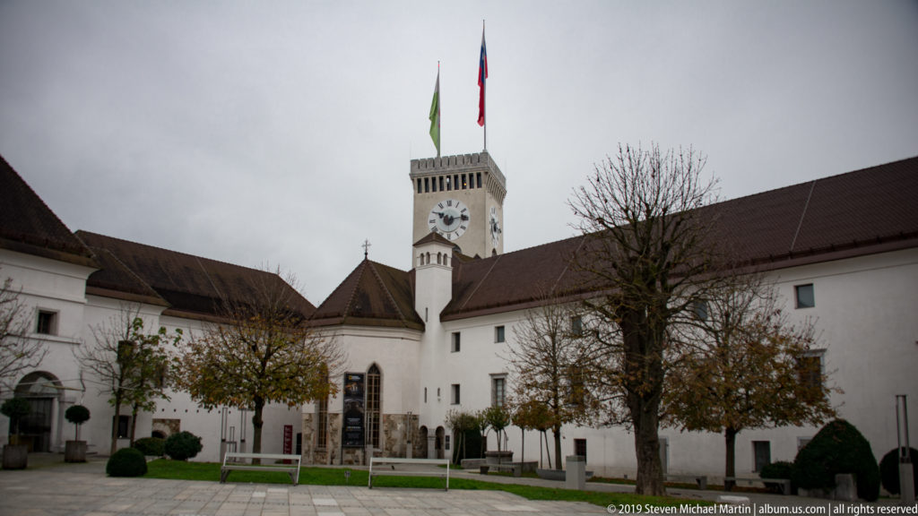 Wide view of Ljubljana Castle courtyard with long white wings, autumn trees, lawn, and the clock tower with flags.