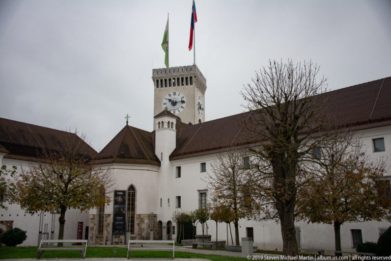 Ljubljana Castle courtyard with white walls, tiled roofs, trimmed trees, flags, and the square clock tower.