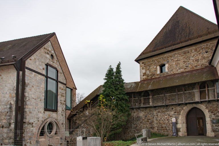 Historic stone buildings at Ljubljana Castle with a glazed opening, timber-covered gallery, arched doorway, and tiled roofs.
