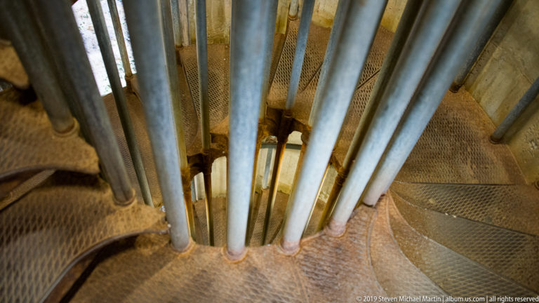 Top-down view of a metal spiral staircase with tubular supports and perforated wedge-shaped steps inside Ljubljana Castle.