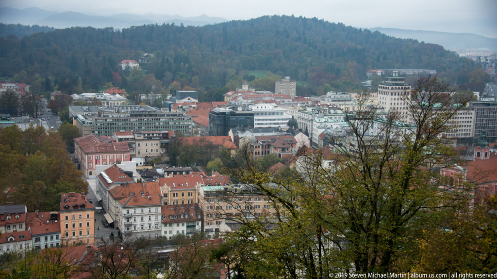 Elevated Ljubljana cityscape with red-roofed buildings, modern blocks, autumn trees, and a wooded ridge in the distance.