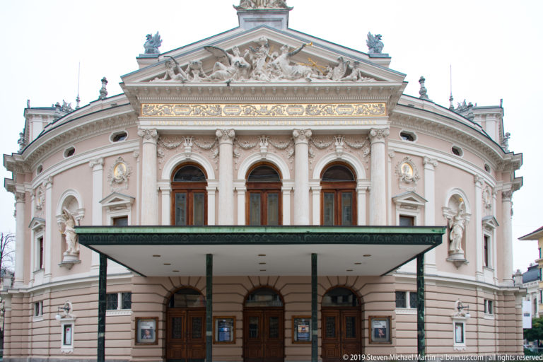 Front entrance of Ljubljana’s pink-and-cream opera house with broad canopy, arched windows, statues, and sculpted pediment.