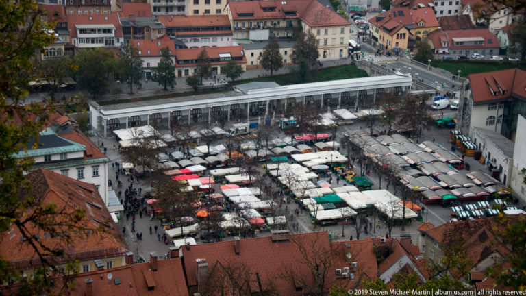 High view over Ljubljana market stalls with rows of white and colored awnings, shoppers, streets, and red-roofed buildings.
