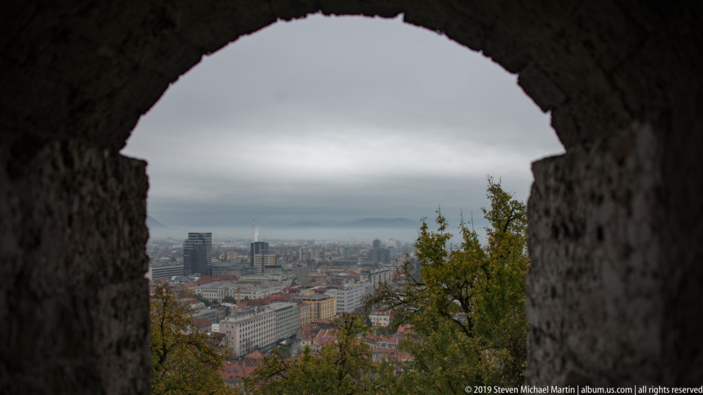 Ljubljana skyline seen through a rough stone opening in Ljubljana Castle, with trees and rooftops beyond.