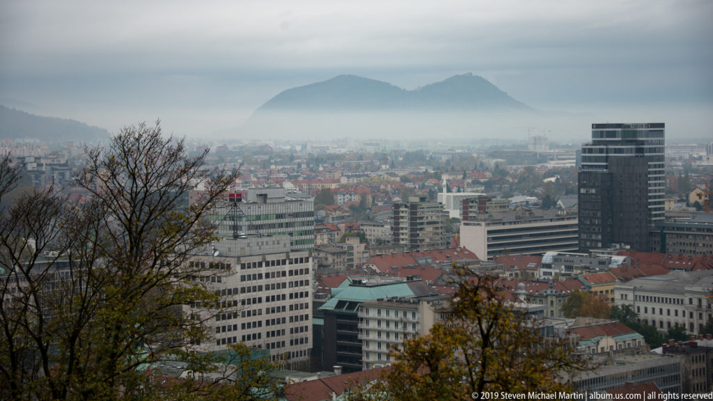 Elevated view over central Ljubljana with red roofs, modern buildings, a dark tower, and hazy hills in the distance.