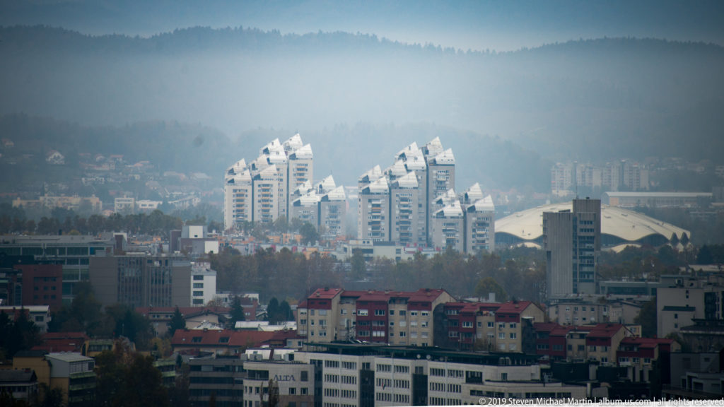 City panorama from Ljubljana Castle toward white high-rise towers, red-roofed neighborhoods, and hazy wooded hills.