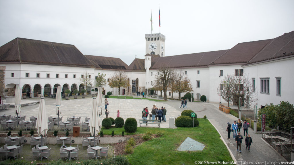 Ljubljana Castle courtyard with white buildings, arcades, outdoor seating, visitors, and a square clock tower.
