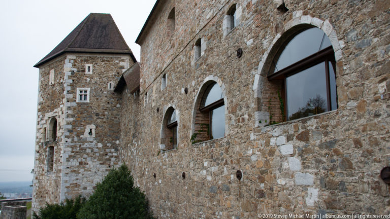Stone exterior of Ljubljana Castle with a projecting tower, rough masonry walls, and large arched glazed openings.