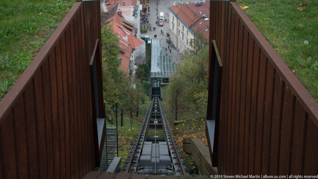 Steep Ljubljana Castle funicular track descending through trees toward rooftops and streets of the city below.