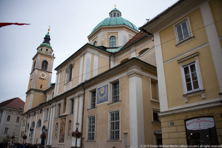 Oblique exterior view of Ljubljana Cathedral with pale Baroque walls, green copper dome, and bell tower.