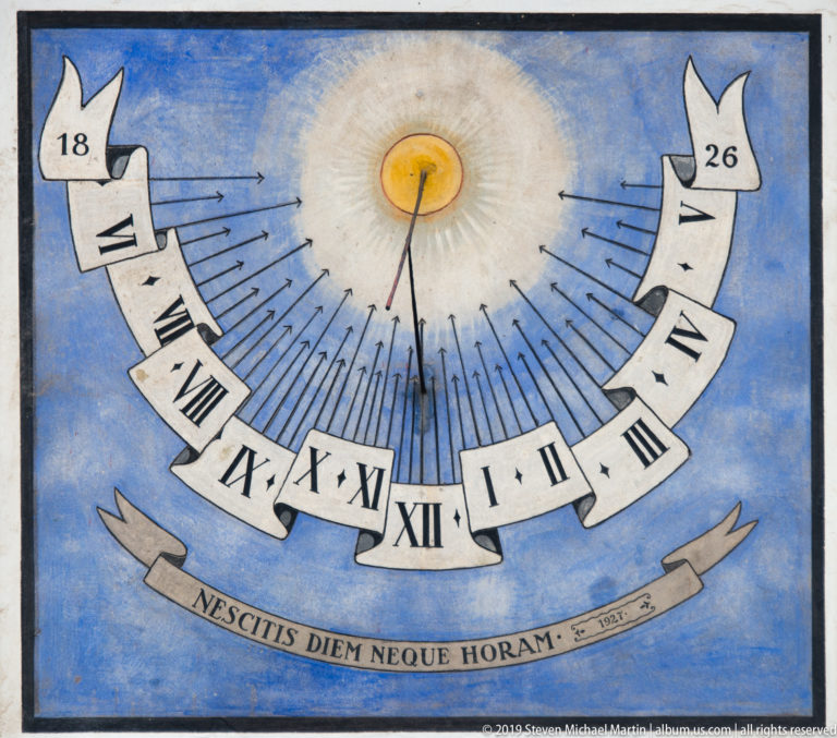 Blue painted sundial with Roman numerals, radiating hour lines, and the Latin motto NESCITIS DIEM NEQUE HORAM.
