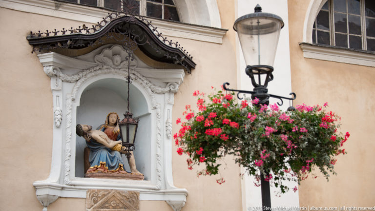 Pietà sculpture of the Virgin Mary holding Christ in a decorative wall niche beside flowers and a streetlamp.