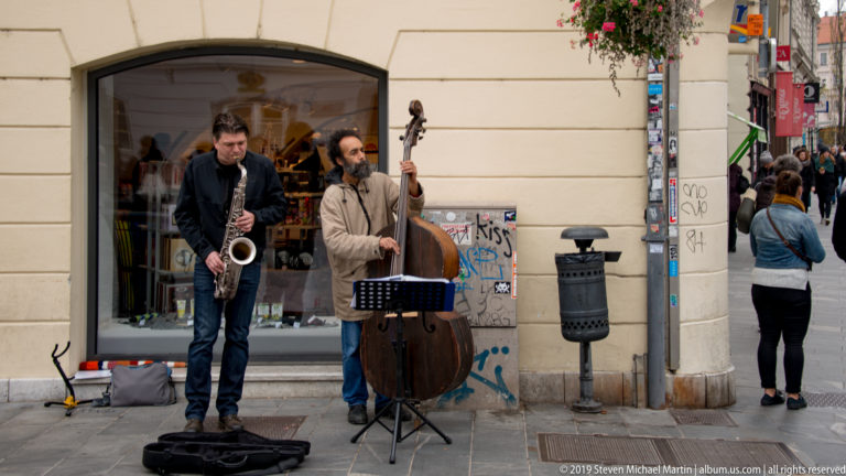 Street saxophonist and double-bass player performing beside a music stand on a pedestrian street in Ljubljana.
