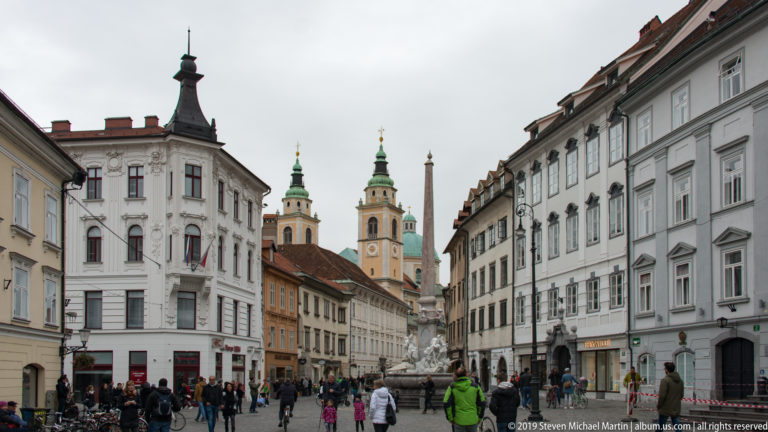 Crowded historic Ljubljana street leading toward the twin bell towers of Ljubljana Cathedral, with a stone monument near the center.