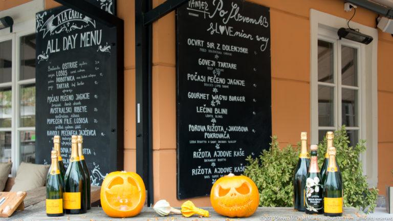 Carved pumpkins, wine bottles, herbs, and blackboard menus arranged outside an orange-painted Ljubljana restaurant.