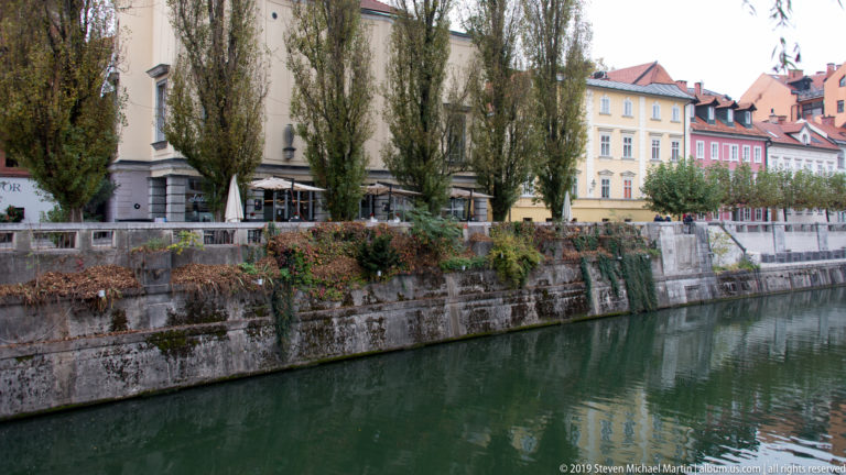 Ljubljanica River beside a stone embankment lined with tall trees, cafés, flowers, and pastel buildings.