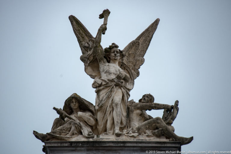 Winged female figure rising above reclining sculpted figures at the roofline of Ljubljana’s opera house.