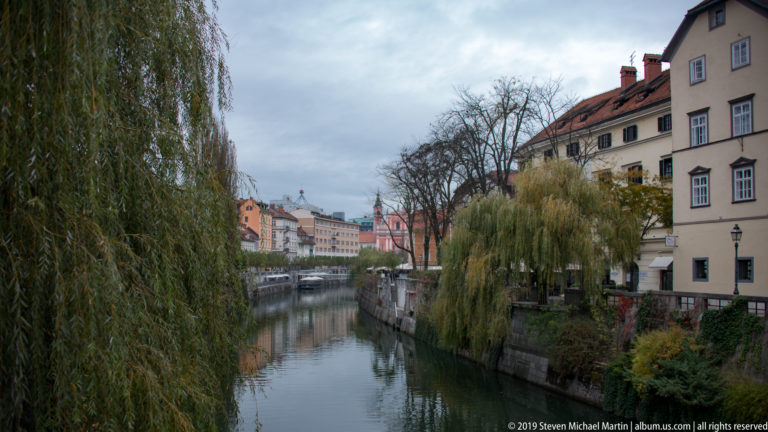 Narrow Ljubljanica River bordered by stone walls, willow trees, and historic buildings under an overcast sky.