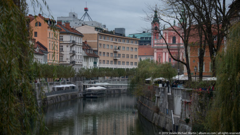 Calm Ljubljanica River bordered by stone embankments, willow trees, historic buildings, and the pink Franciscan church in the distance.