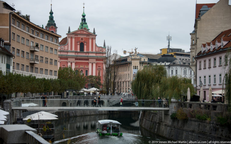 Ljubljanica River with a small tour boat, riverside cafés, bridge, and the pink Franciscan Church of the Annunciation beyond.