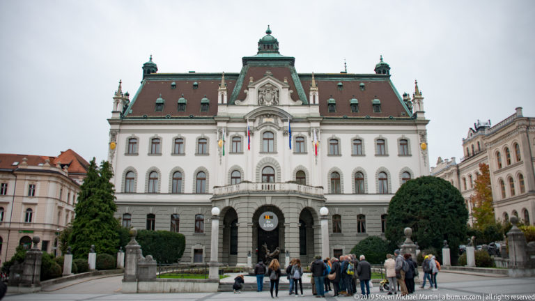 University of Ljubljana main building with symmetrical white façade, steep green roof, central entrance, and people gathered outside.