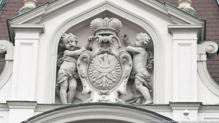 Crowned heraldic shield in pale stone flanked by two sculpted putti within an ornate pediment.