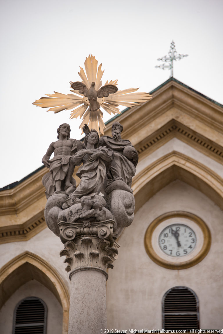 Stone religious figures on a carved column capital beneath a golden dove and rays, with a church clock nearby.