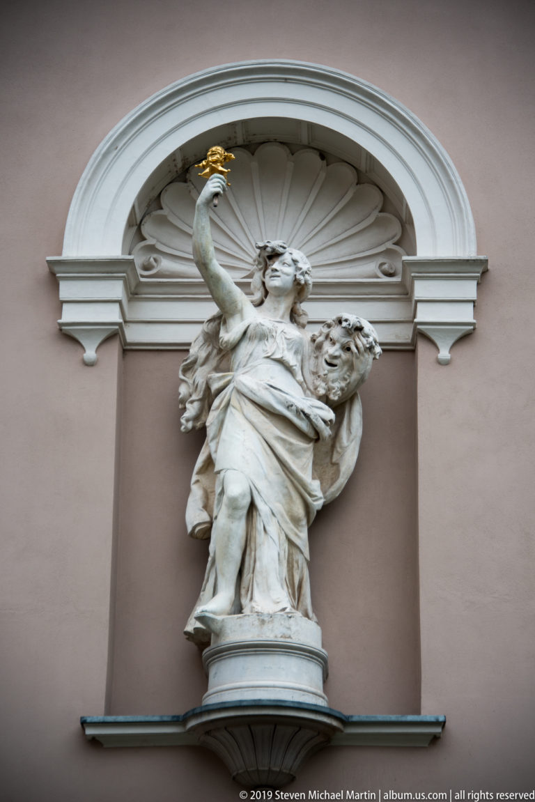 White allegorical female statue raising a gilded object in an arched niche on Ljubljana’s opera house.