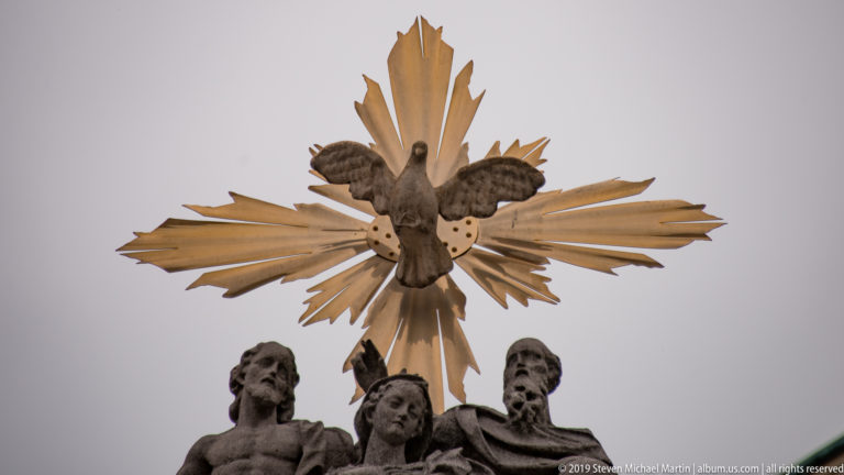 Golden dove and radiating rays above dark sculpted religious figures on a Ljubljana monument.