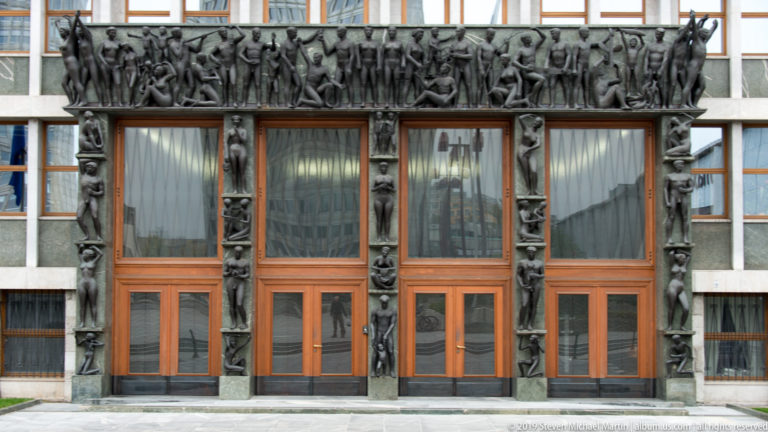 National Assembly entrance in Ljubljana with bronze relief figures surrounding four tall glazed wooden doors.