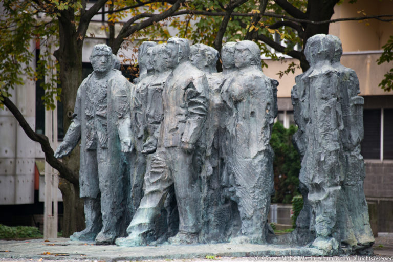 Cluster of rough-textured bronze human figures standing closely together in an outdoor memorial sculpture.
