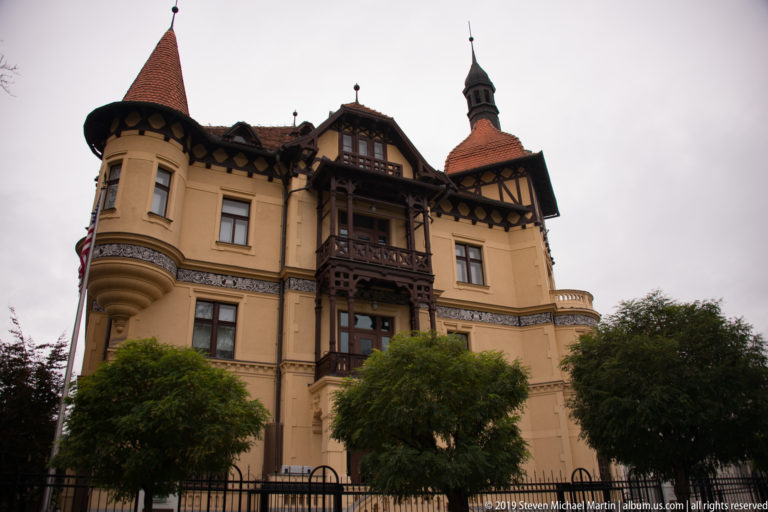 Ornate yellow historic villa with timber balconies, rounded bays, decorative friezes, and a narrow turret behind trees.