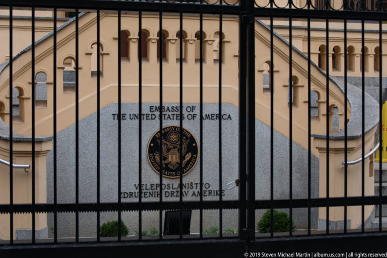 United States Embassy sign and seal in Ljubljana seen through a black metal security fence.