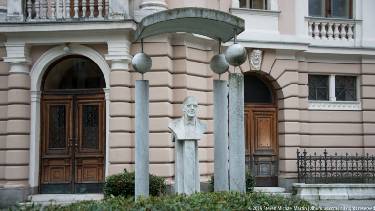 Stone portrait bust between slender columns beneath a curved metal canopy in front of a historic building.