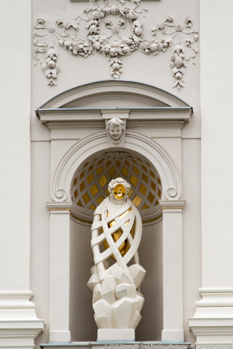Close view of a pale figure wrapped by interlacing sculptural bands beneath a gilded accent on the National Gallery.
