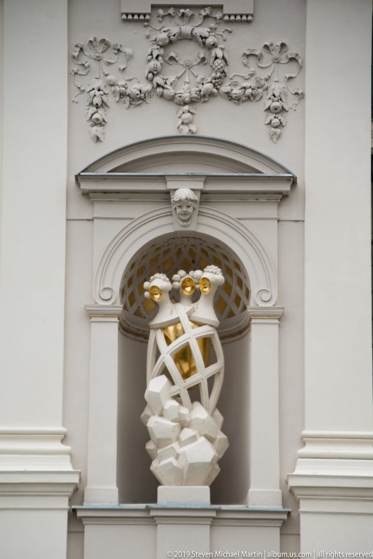 White sculptural figure enclosed by interlacing bands and gilded details in a niche on the National Gallery façade.