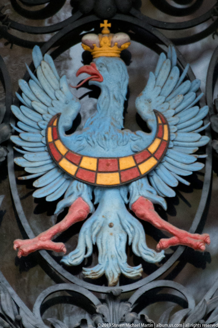 Close-up of a painted crowned eagle with pale blue wings, red beak and legs, gold crown, and red-and-gold band.