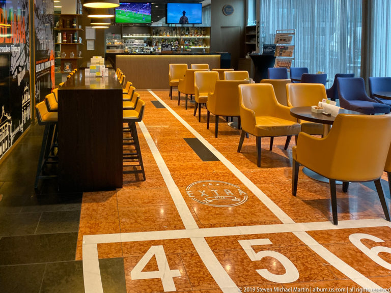 Contemporary hotel lounge with numbered running-track graphics on the polished floor, yellow chairs, communal table, and bar.