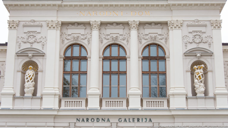Ornate upper façade of the National Gallery of Slovenia with arched windows, columns, statues, and Narodna galerija inscription.