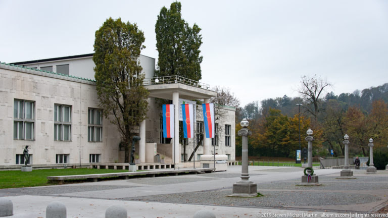 Stone modernist façade of Ljubljana’s Moderna galerija with Slovenian flags beside the entrance and trees beyond.