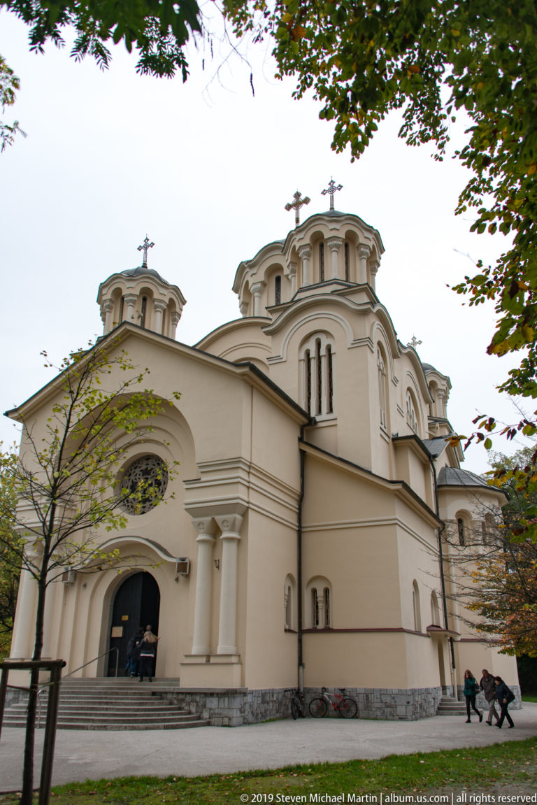 Cream-colored Church of Saints Cyril and Methodius with rounded gables, arched entrance, and multiple crosses among trees.