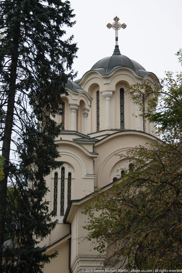 Upper portion of the Church of Saints Cyril and Methodius with rounded gables, dark dome, and Orthodox cross behind trees.